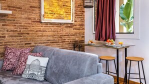 Ein gemütliches Wohnzimmer mit grauem Sofa, gemusterten Kissen, einem gerahmten Bild, einem kleinen Tisch mit Früchten und Getränken und einem Fenster mit Vorhang.