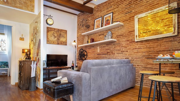 Ein gemütliches Wohnzimmer mit Backsteinwand, einem grauen Sofa, einem Fernseher und einer kleinen Essecke mit Barhocker.