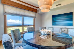 A modern dining area with a round glass table, grey upholstered chairs, and a large abstract painting on the wall. - The Resort at Kapalua Bay | Ocean View 3 BR | Car Incl. w/ 6+ Nights | MON-6401 by KBM (Lahaina)