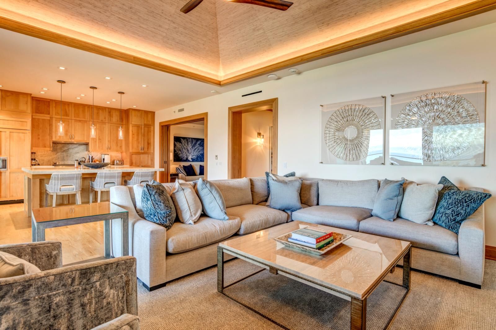 Uma sala de estar moderna com um amplo sofá modular, uma mesa de centro de vidro e uma área de cozinha com arm ários de madeira e iluminação pendente.