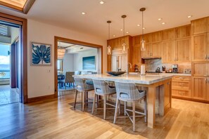 A modern kitchen with wooden cabinets, a large island with bar stools, pendant lights, and a view to the dining area.