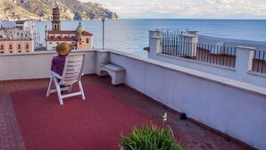 A balcony with a view of the sea and a mountain, a person sitting on a chair, and a potted plant.
