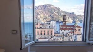 A room with a large window offering a view of a coastal town, a table with lemons, and a bottle.