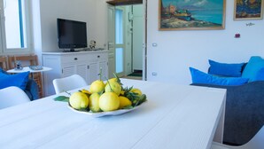 A modern living room with a white table, a bowl of lemons, a grey sofa with blue cushions, and a painting of a coastal scene on the wall.