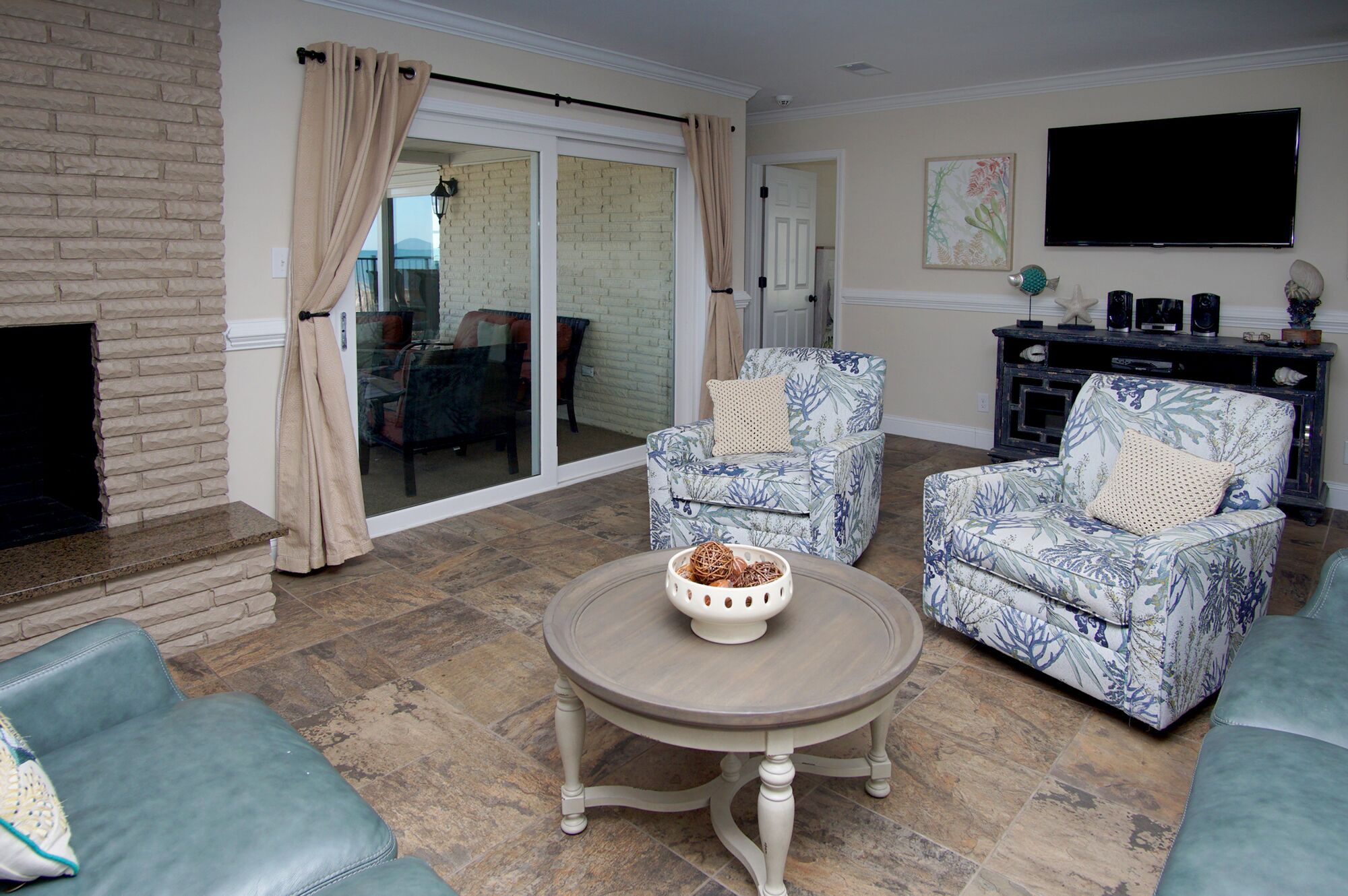 A living room with a fireplace, two patterned armchairs, a round table with a decorative bowl, and a television mounted on the wall.