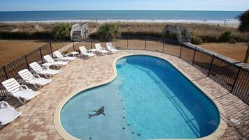 A swimming pool with a view of the beach, surrounded by white lounge chairs and a brick deck.