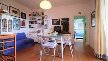 A cozy living room with a blue sofa, a glass table, and metal chairs. There are shelves with books and decorative items, and a TV on a stand.