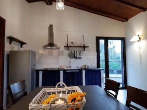A kitchen with a stove, sink, and hanging utensils. There is a dining table with chairs and a basket of flowers. - Casa La Violara (Santa Margherita Ligure)