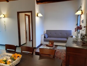 A living room with a grey sofa, wooden chairs, a coffee table, and a dining table. There are wall-mounted lamps and a vase with flowers on the coffee table.