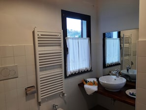 A bathroom with a white tiled wall, a white towel rack, a window with a white curtain, a mirror, a sink, and a towel with a floral arrangement.