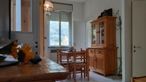 A dining area with a wooden table and chairs, a cabinet with glass doors, and a view of greenery through a window. - Casa Isotta (Bonassola)