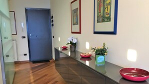 A modern hallway with a wooden floor, a glass partition, and a door. There are two framed artworks on the wall and a vase with flowers on the counter.