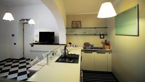 Cozinha compacta com fogão a gás, micro-ondas e uma pequena pia. Há uma mesa de jantar com cadeiras, piso xadrez preto e branco e luminárias pendentes.