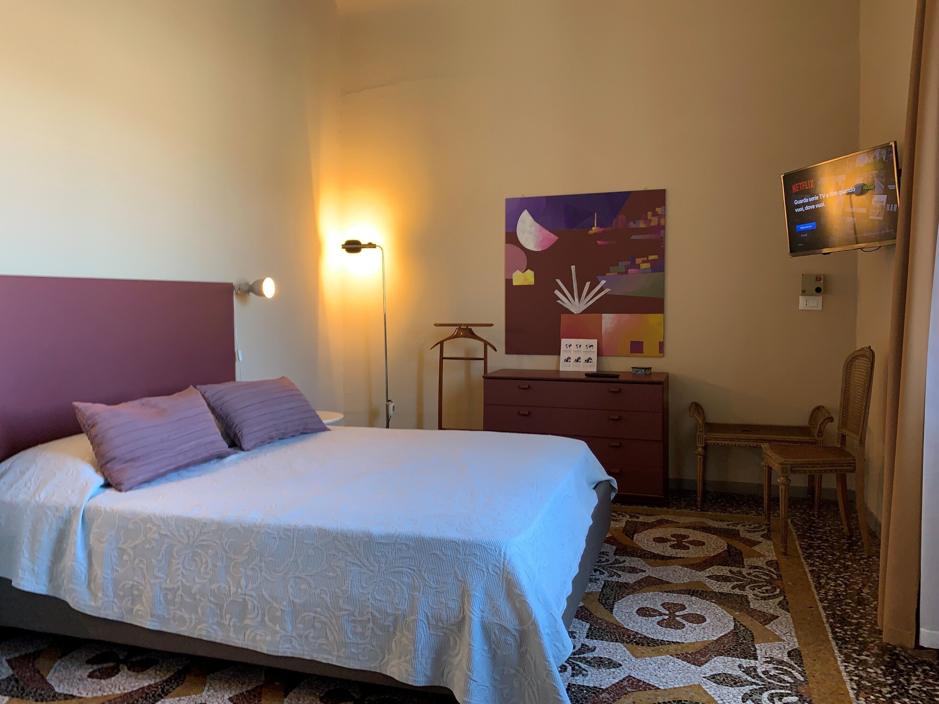 Quarto de hotel com cama, escrivaninha, cadeira, luminária, um quadro e televisão na parede.
