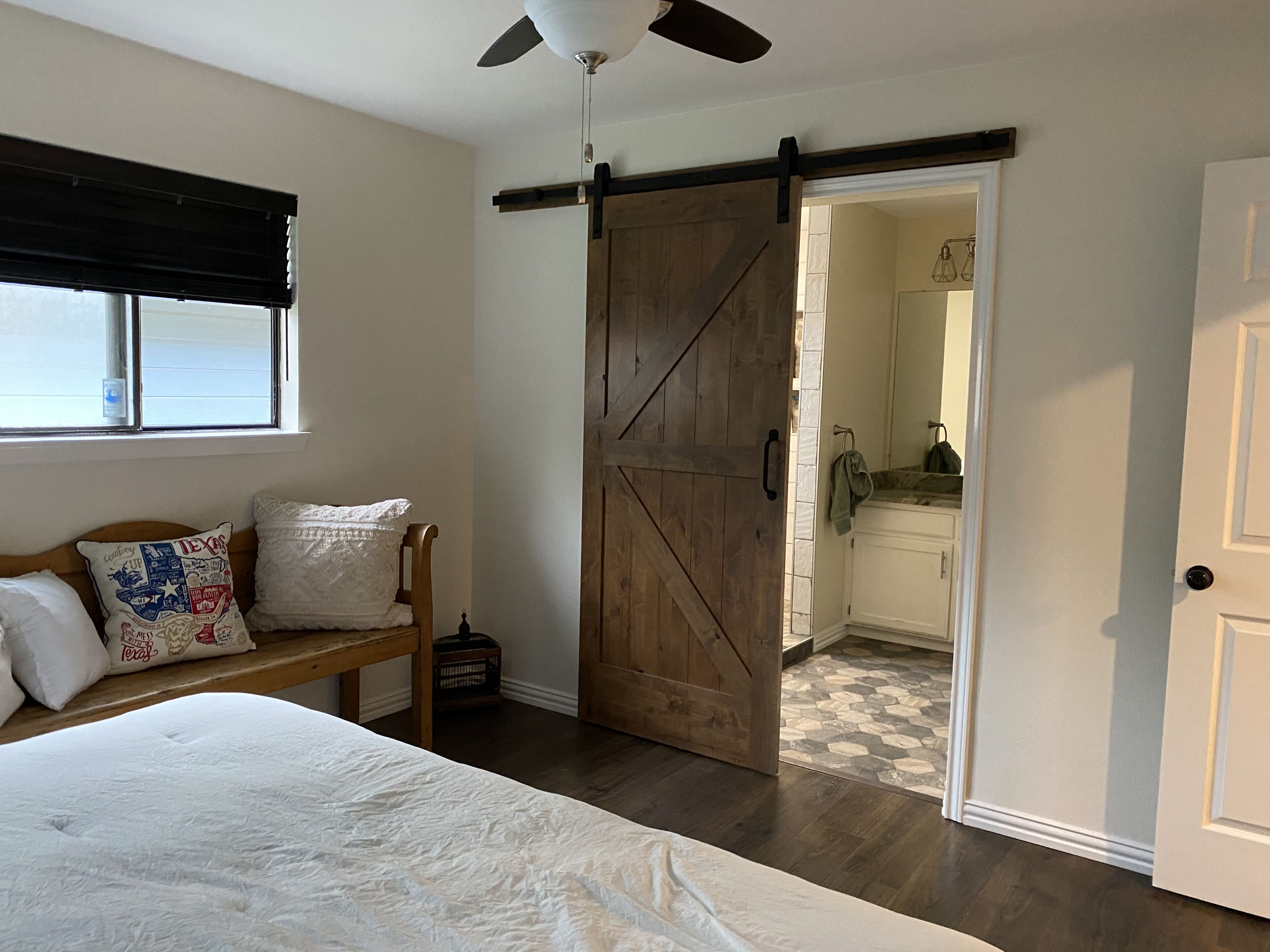 A bedroom with a bed, a bench with pillows, a wooden door, and a view into a bathroom.
