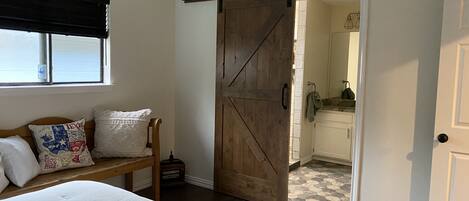 A bedroom with a bed, a bench with pillows, a wooden door, and a view into a bathroom.