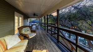 A covered porch with a wooden deck, a wicker chair, a striped sofa, and a view of trees and a building in the background. - Picturesque and Beautiful 3B on the Guadalupe River (New Braunfels)