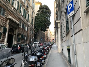 Une rue bordée de voitures et de motos garées, un panneau de signalisation indiquant un espace de stationnement et des bâtiments à l’architecture classique.