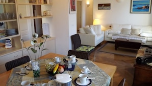 Comedor con mesa lista para dos, cafetera y un florero con flores. Se ve un sofá y una lámpara al fondo.