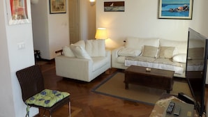 Una sala de estar con un sofá blanco, una mesa de centro de madera, una silla con cojín floral, un televisor y obras de arte en las paredes.