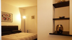 A bedroom with a bed, a nightstand, a floor lamp, and a wall-mounted shelf with decorative items. - Casa Ninni (Chiavari)