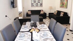 A dining area with a table set for four, surrounded by chairs, with a black sofa and a painting in the background. - Casa Ninni (Chiavari)