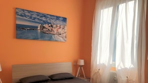 A bedroom with a bed, a nightstand, a lamp, a window with sheer curtains, and a painting on the wall. - House Nervi (Genoa)
