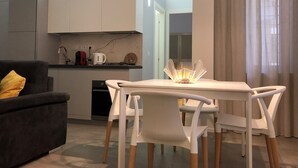 Una cocina moderna con armarios blancos, estufa a gas y cafetera. Hay una mesa de comedor con sillas blancas y un candelabro de vidrio.