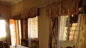 A dining area with tables set for two, wooden chairs with green cushions, and curtains with a floral pattern. - Magline Conference And Guest House (Kisumu)