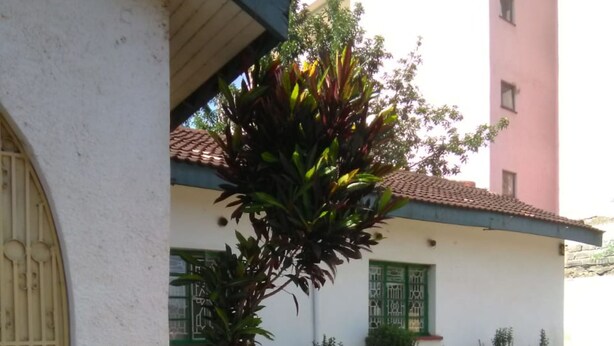 A building with a white exterior, a tiled roof, and a large door. There are steps leading to the entrance, and a small garden area with various plants. - Magline Conference And Guest House (Kisumu)