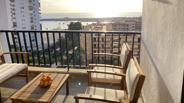 Ein Balkon mit Holzmöbeln, einem Tisch mit Orangen und Blick auf das Meer und Gebäude.