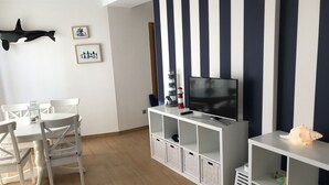 A living room with a white TV stand, a flat-screen TV, a dining table with white chairs, and a wall-mounted shelf with decorative items. - OH LA LIFE! PORTOSIN SAN JOSÉ (Portosin)