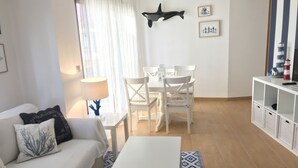A living room with a white sofa, a coffee table, and a dining area with white chairs and a table. There is a wall-mounted decoration and a small cabinet with baskets. - OH LA LIFE! PORTOSIN SAN JOSÉ (Portosin)