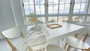 Una habitación con un ventanal grande con vistas a una playa, una mesa blanca con sillas y una manta a rayas en el suelo.