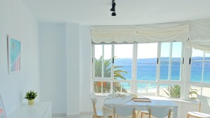 Habitación luminosa con un amplio ventanal con vistas a la playa. Hay un escritorio blanco con una planta, una mesa con sillas y una cama con cubierta blanca.