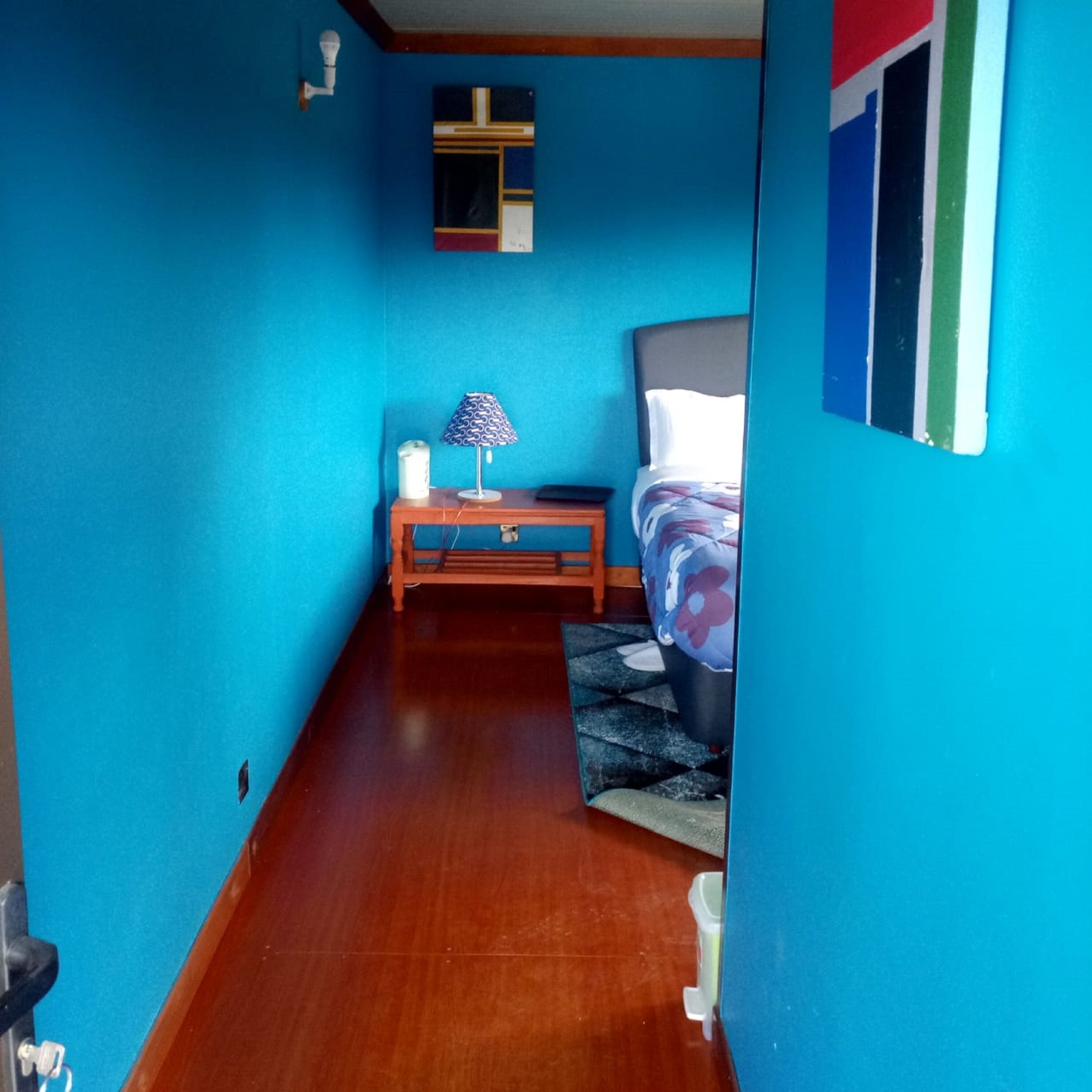 Un couloir étroit mène à une chambre aux murs bleus, dotée d’un plancher en bois, d’une table de chevet avec une lampe et d’un lit recouvert d’une couvre-lit à motifs floraux.
