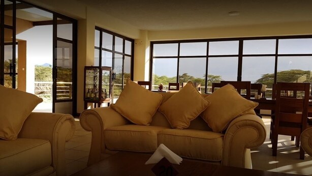 A living room with a beige sofa set, a wooden coffee table, and large windows offering a view of the outdoors. - Matteo's Resort (Naivasha)