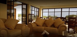 A living room with a beige sofa set, a wooden coffee table, and large windows offering a view of the outdoors. - Matteo's Resort (Naivasha)