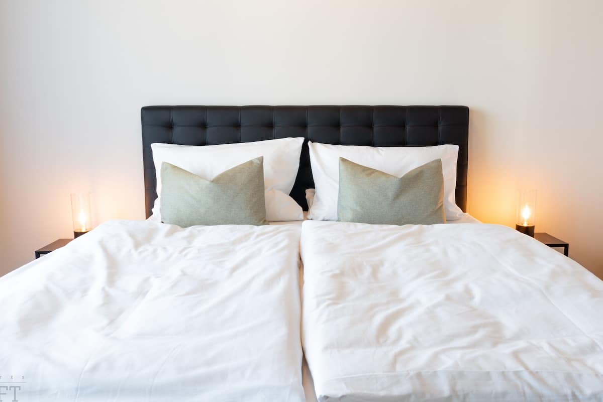 A neatly made bed with white linens and two pillows, a dark headboard, and two bedside lamps.