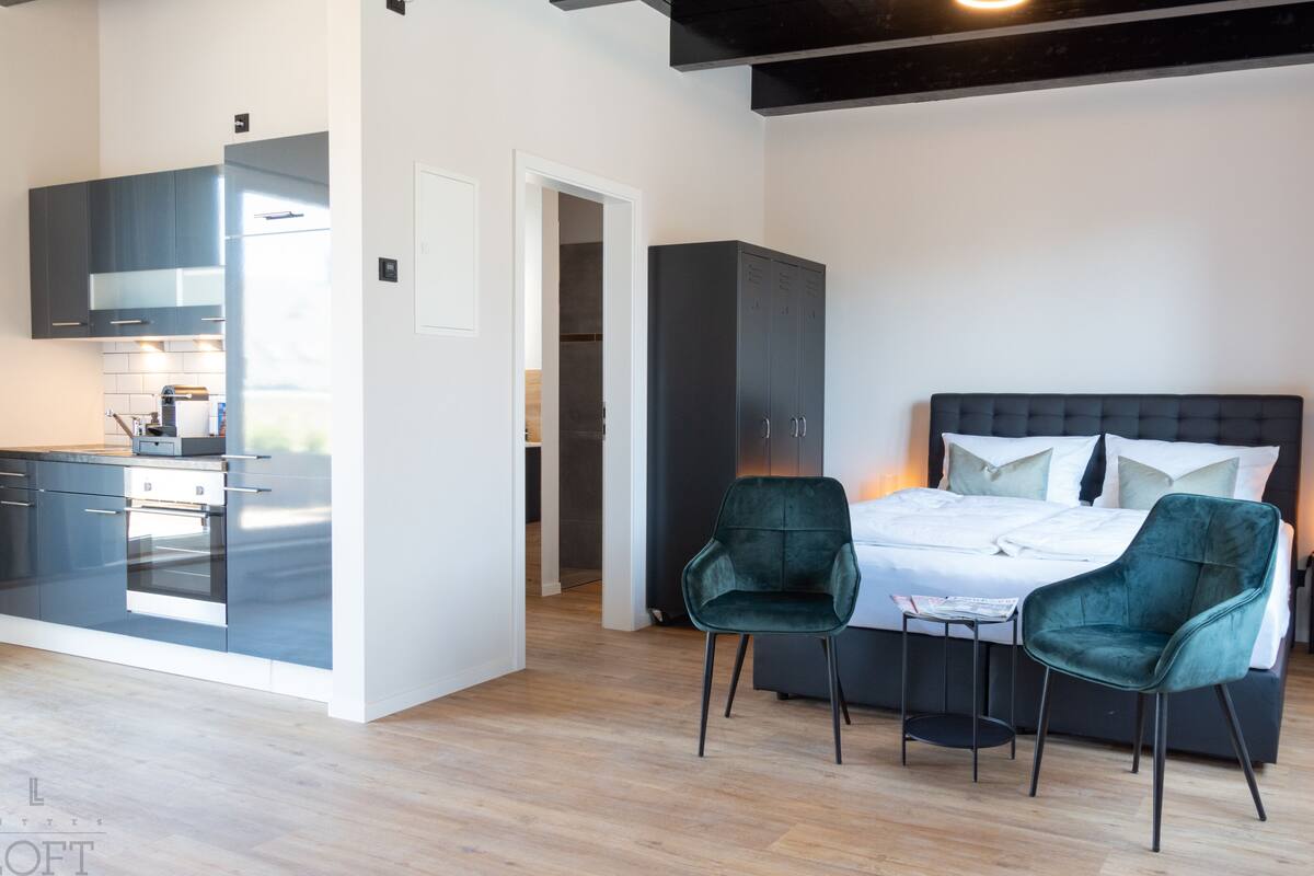 A modern hotel room with a bed, two chairs, a small table, a wardrobe, and a kitchenette.