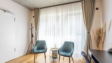 A room with two teal chairs, a small round table, a dark wooden desk, and a large window with sheer curtains.