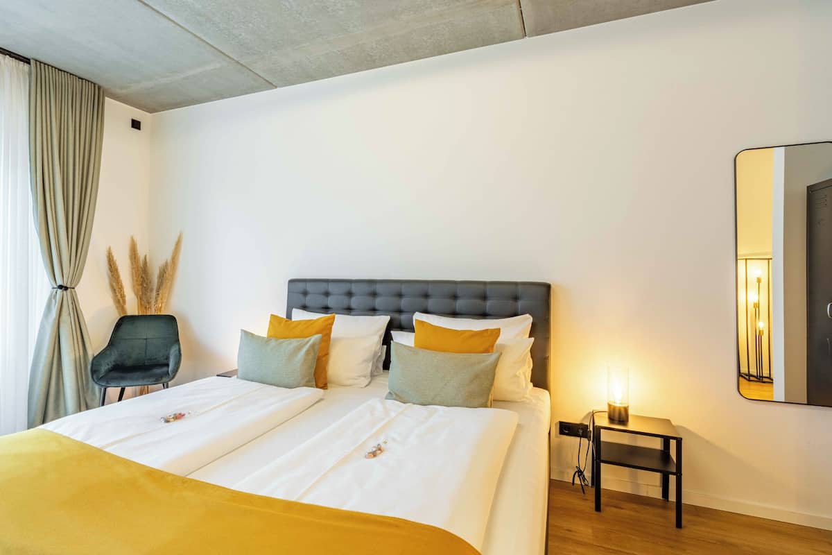 A modern hotel room with a large bed, a nightstand, a chair, and a mirror on the wall.