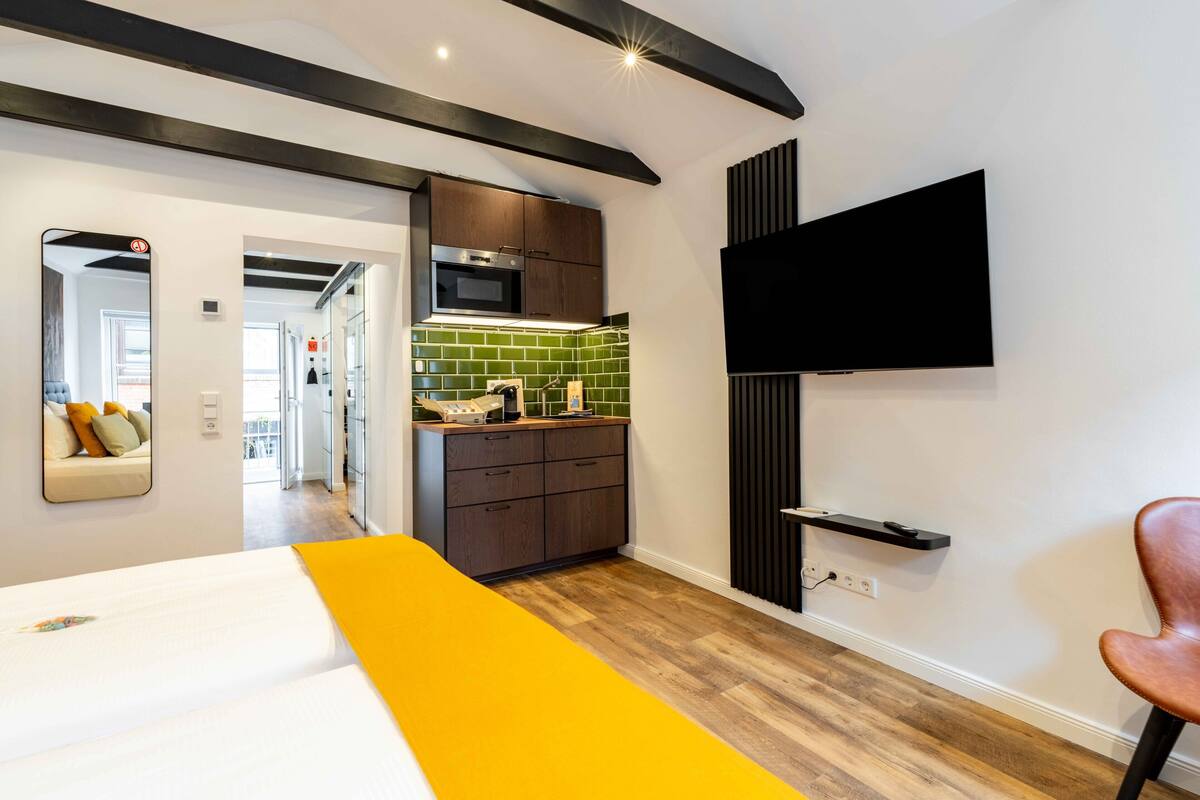A modern hotel room with a bed, a TV mounted on the wall, a kitchen area with a microwave and a sink, and a wooden floor.