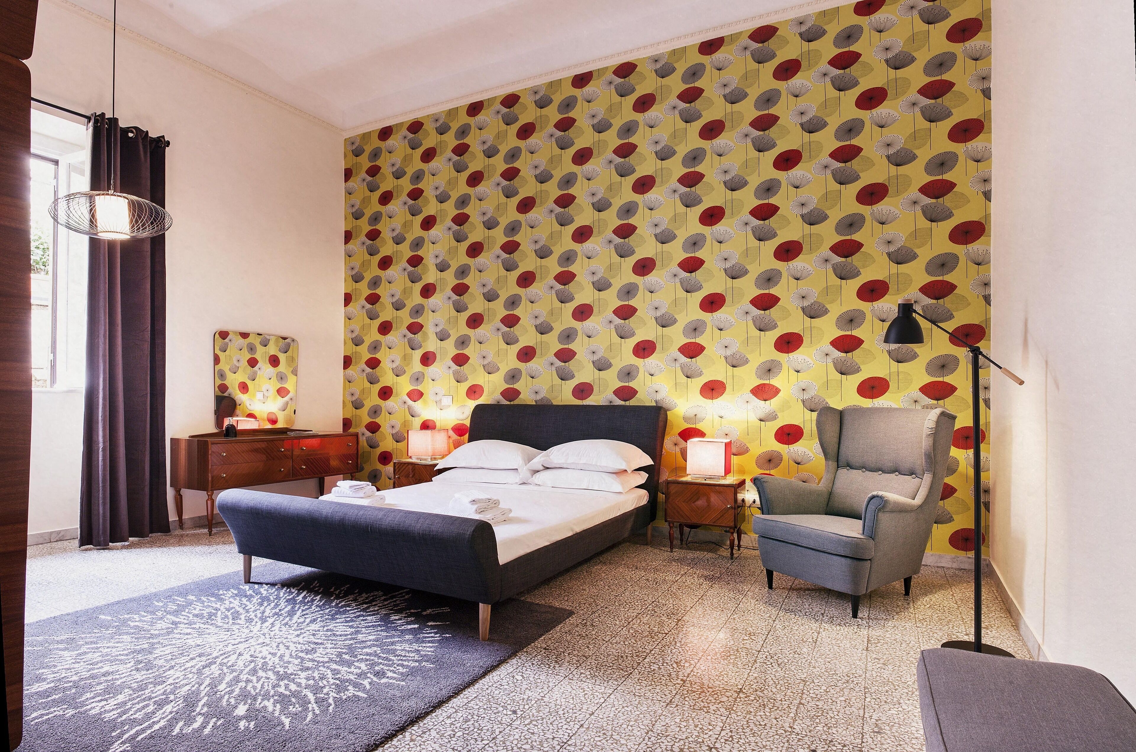 A bedroom with a bed, a chair, a lamp, and a patterned wallpaper.