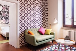 Una habitación con un sillón verde, una mesita blanca, un papel tapiz estampado y un radiador.