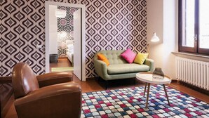 Un salon moderne avec un papier peint à motifs, un canapé vert agrémenté de coussins colorés, un fauteuil en cuir marron, une petite table blanche et un radiateur.