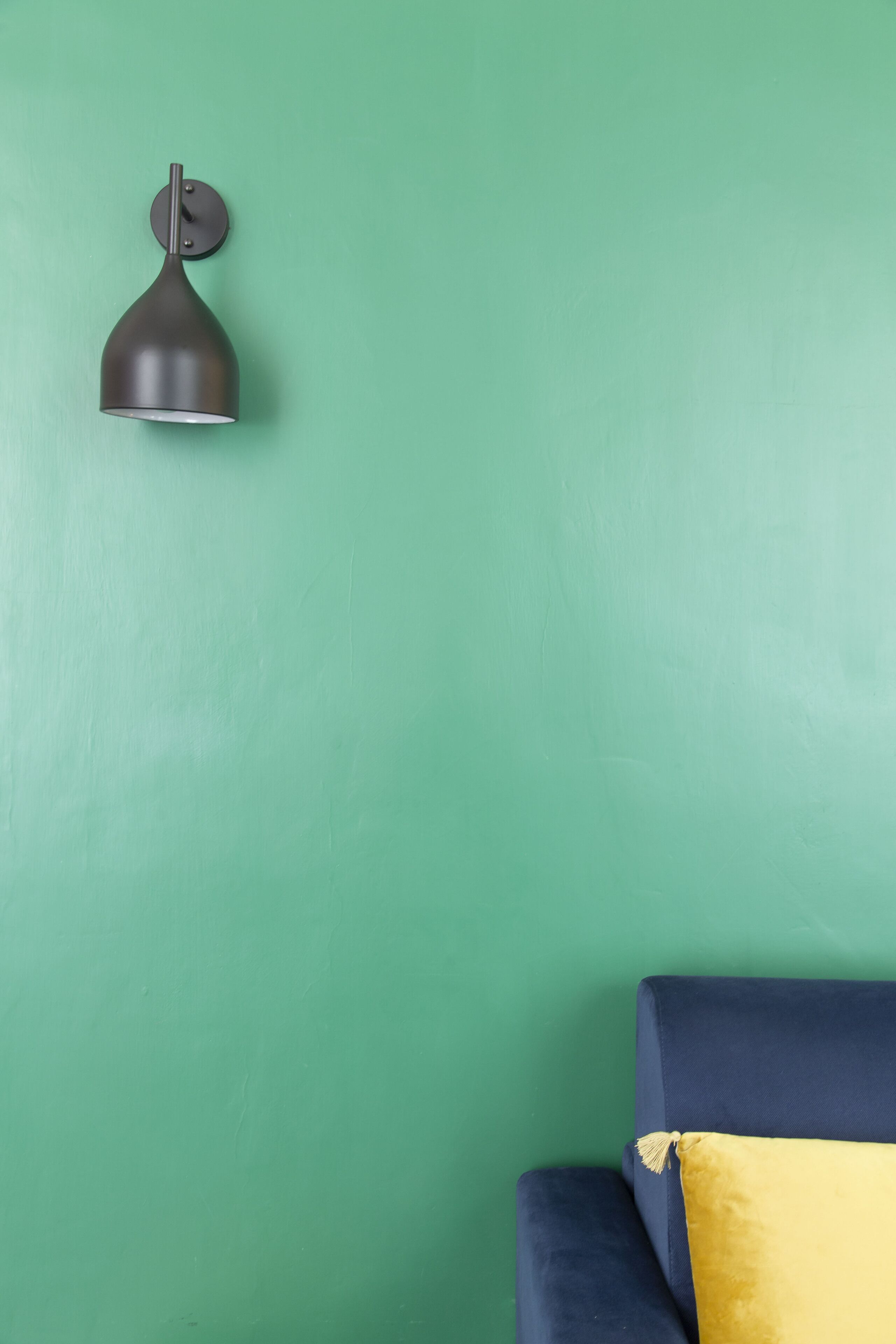 A green wall with a dark pendant light fixture and a dark blue sofa with a yellow pillow.