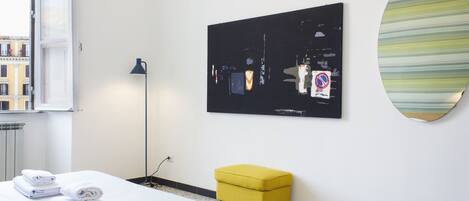 A hotel room with a bed, a yellow ottoman, a black artwork, and a round mirror on the wall.