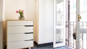 Eine weiße Kommode mit Schubladen, eine Vase mit rosa Blumen und eine offene Tür zu einem Balkon mit Geländer und einer hängenden Pflanze.