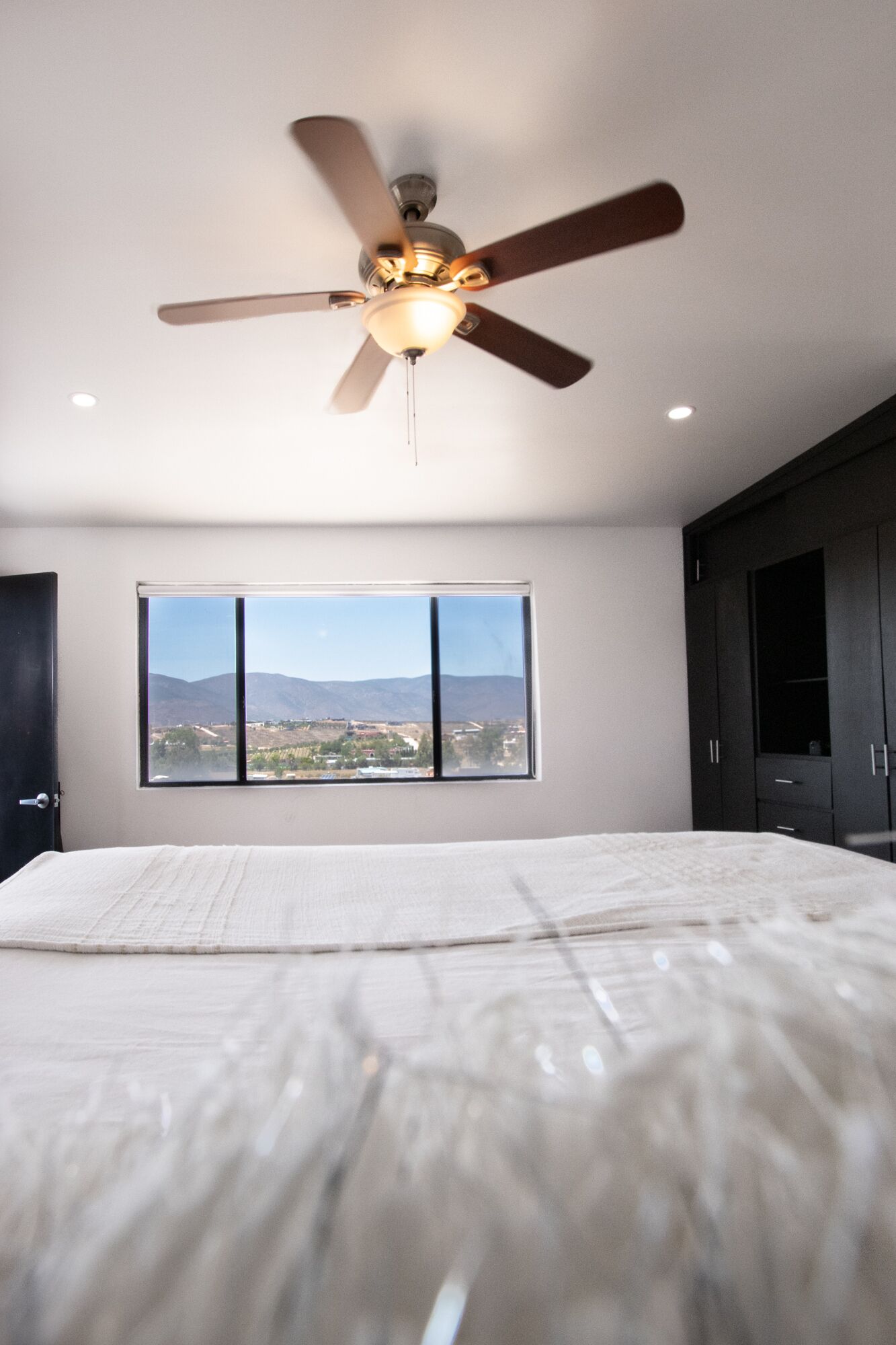 A bedroom with a bed, a ceiling fan, a window with a mountain view, and dark wooden cabinets.